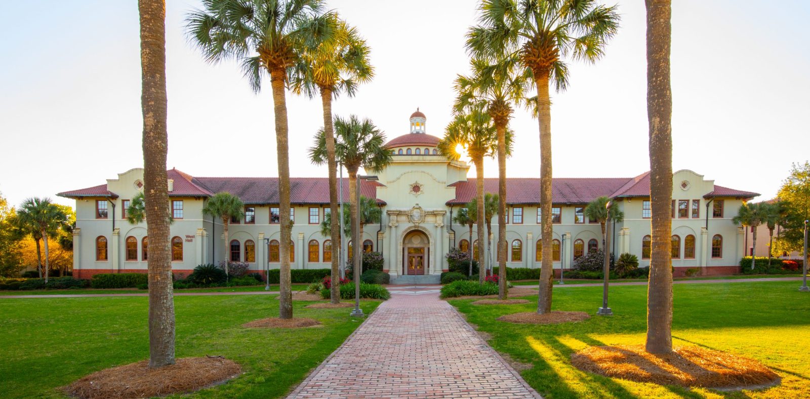 Big two story light colored building with red roof and palm trees.